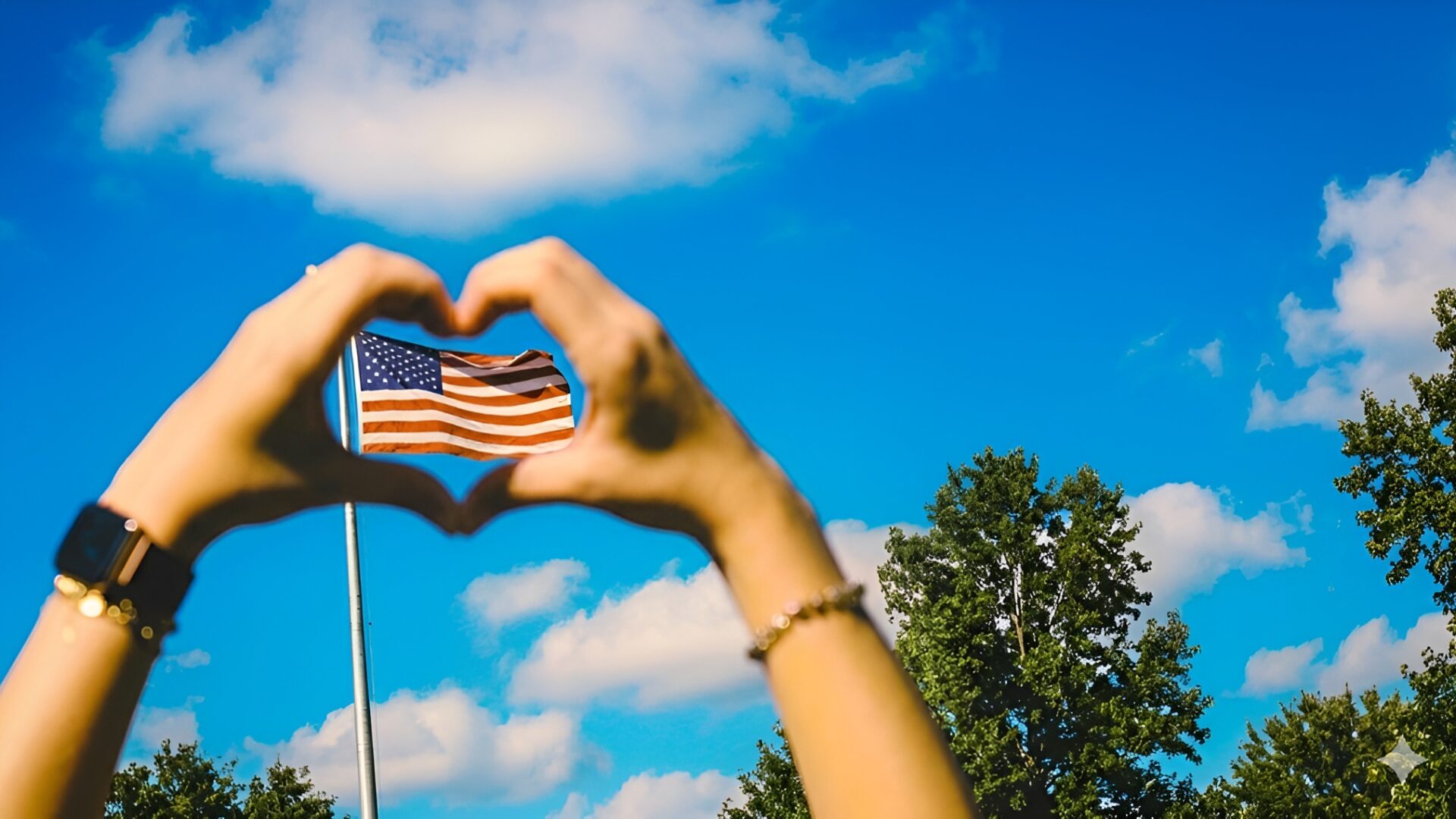 Hands forming a heart over a waving american flag in a blue sky